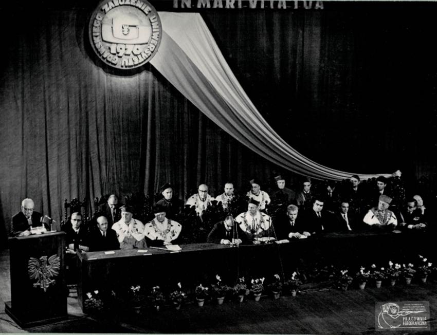 Rektor, dziekani i uroczyście ubrani dygnitarze siedzą za długim stołem na scenie na tle kotary. Przy mównicy stoi jeden z mężczyzn w garniturach. 