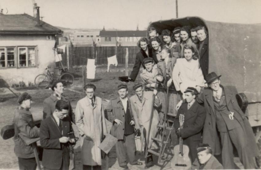 Grupa uśmiechniętych młodych kobiet i mężczyzn w czapkach studenckich przy ciężarówce, która stoi na wiejskim podwórku. 