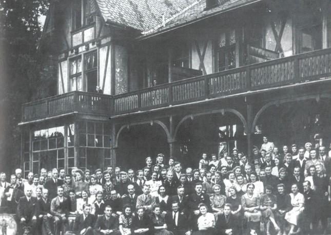 Grupowe zdjęcie studentek, studentów i nauczycieli, pozujących przed piętrowym budynkiem z drewnianym tarasem i balkonami. 