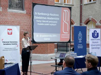 Otwarcie konferencji na dziedzińcu Politechniki Gdańskiej. Kobieta z mikrofonem stoi przed ekranem z prezentacją.